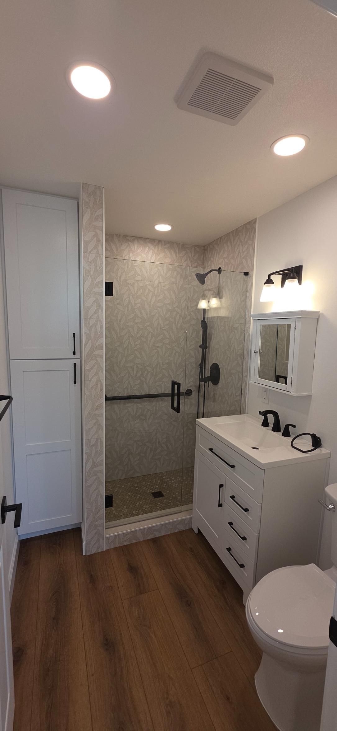 Bright bathroom with glass shower, white vanity, toilet, and wood-look floor.