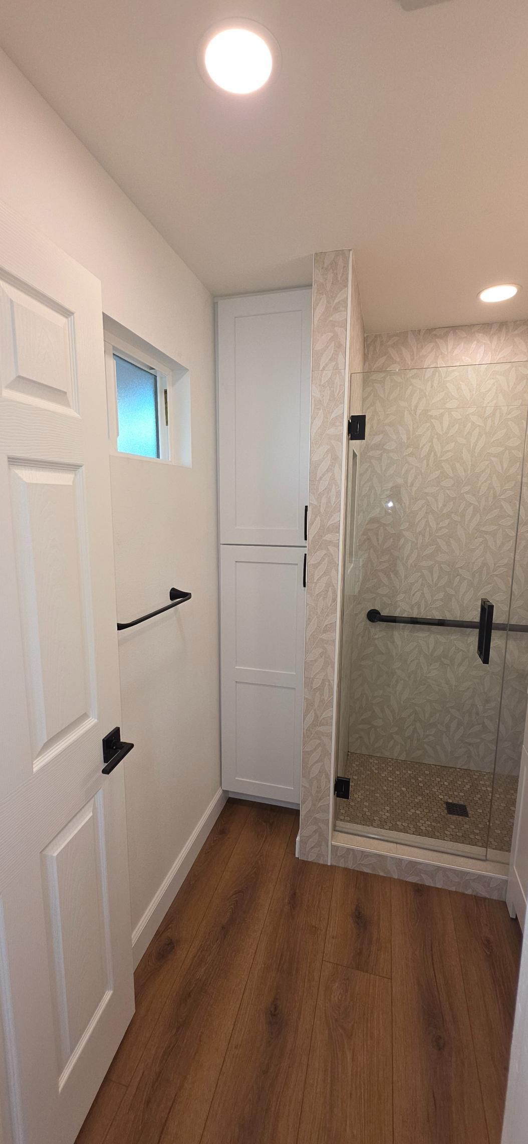 Narrow bathroom with white walls, wood floor, and glass shower enclosure.
