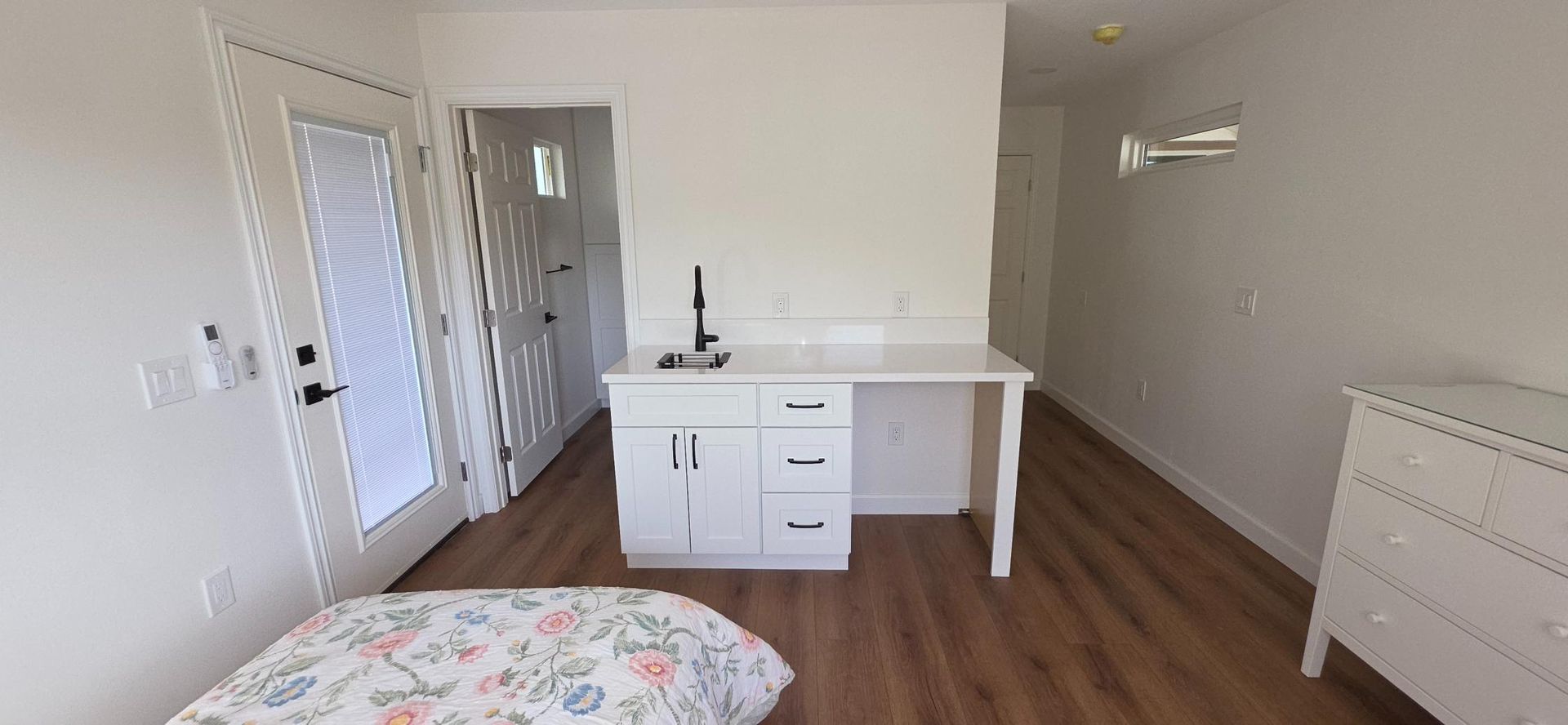 Bright bedroom with white walls, wood floors, bed, closet doors, and built-in dresser by a window