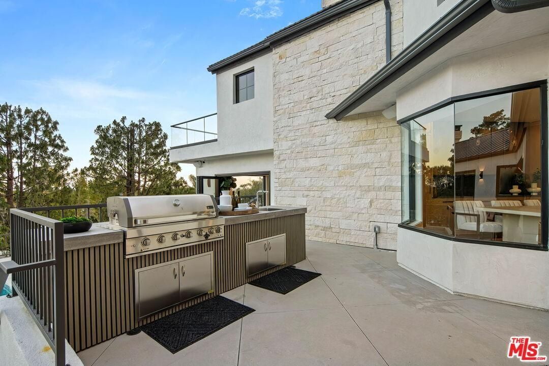 Modern patio with outdoor kitchen and grill beside a stone house and large windows