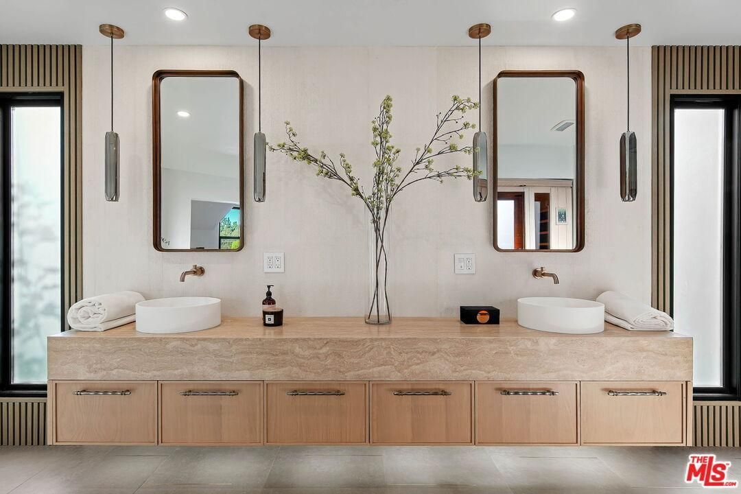 Modern double vanity bathroom with light wood cabinetry, marble countertop, mirrors, and floral arrangement.