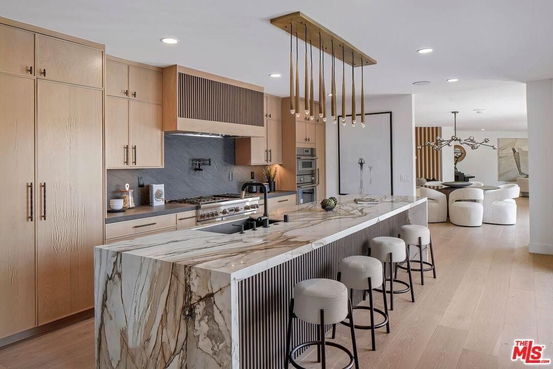 Bright modern kitchen with marble island, wood cabinetry, bar stools, and a sitting area beyond.
