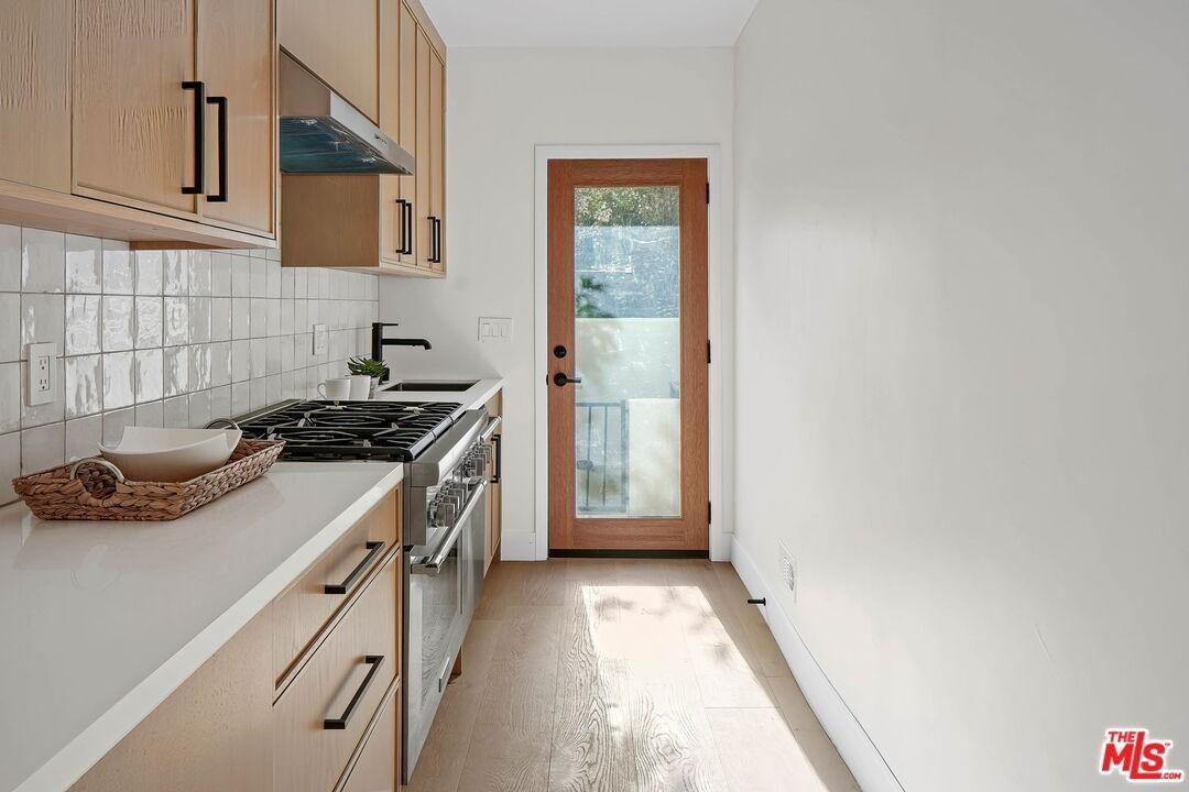 Narrow modern kitchen with wood cabinets, white countertops, stainless stove, and a glass door at the end.