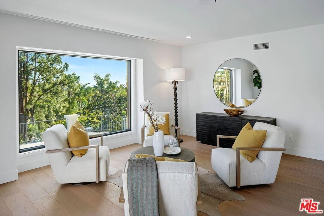 Bright living room with white chairs, yellow pillows, large window, round mirror, and city view