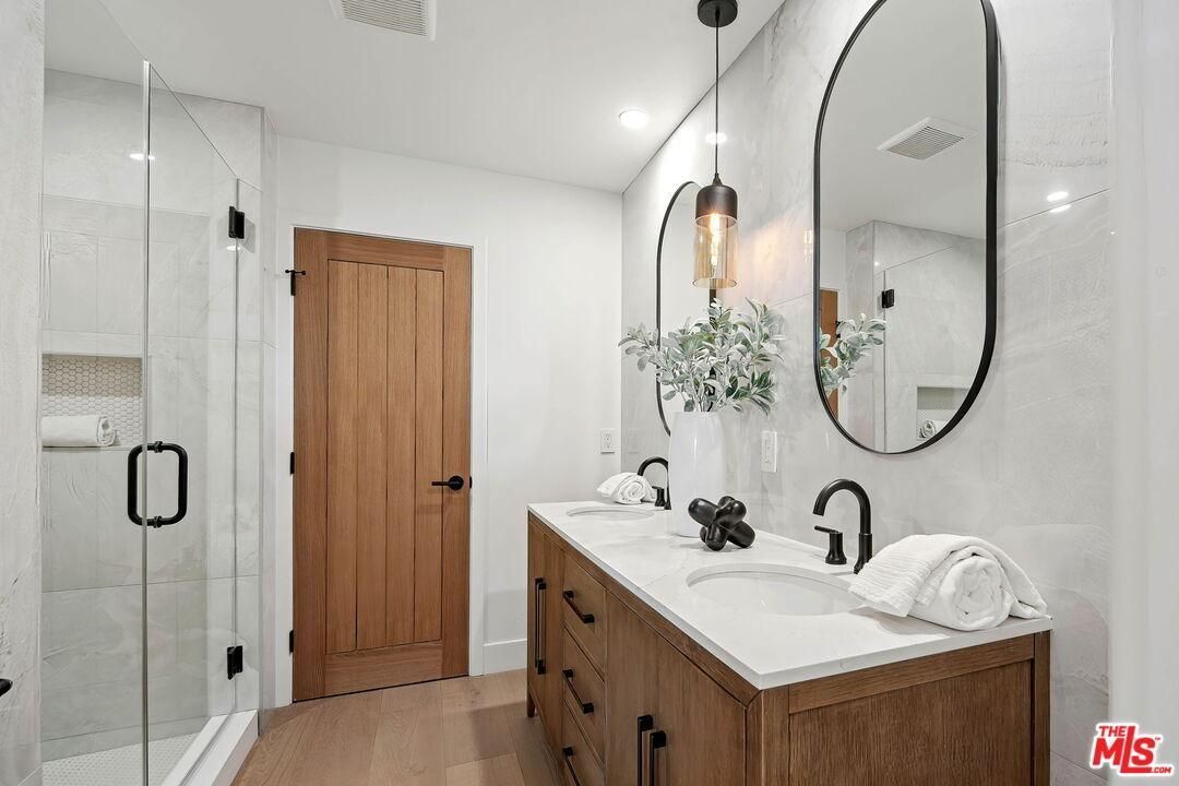 Modern bathroom with wood vanity, round mirror, glass shower, and white walls.