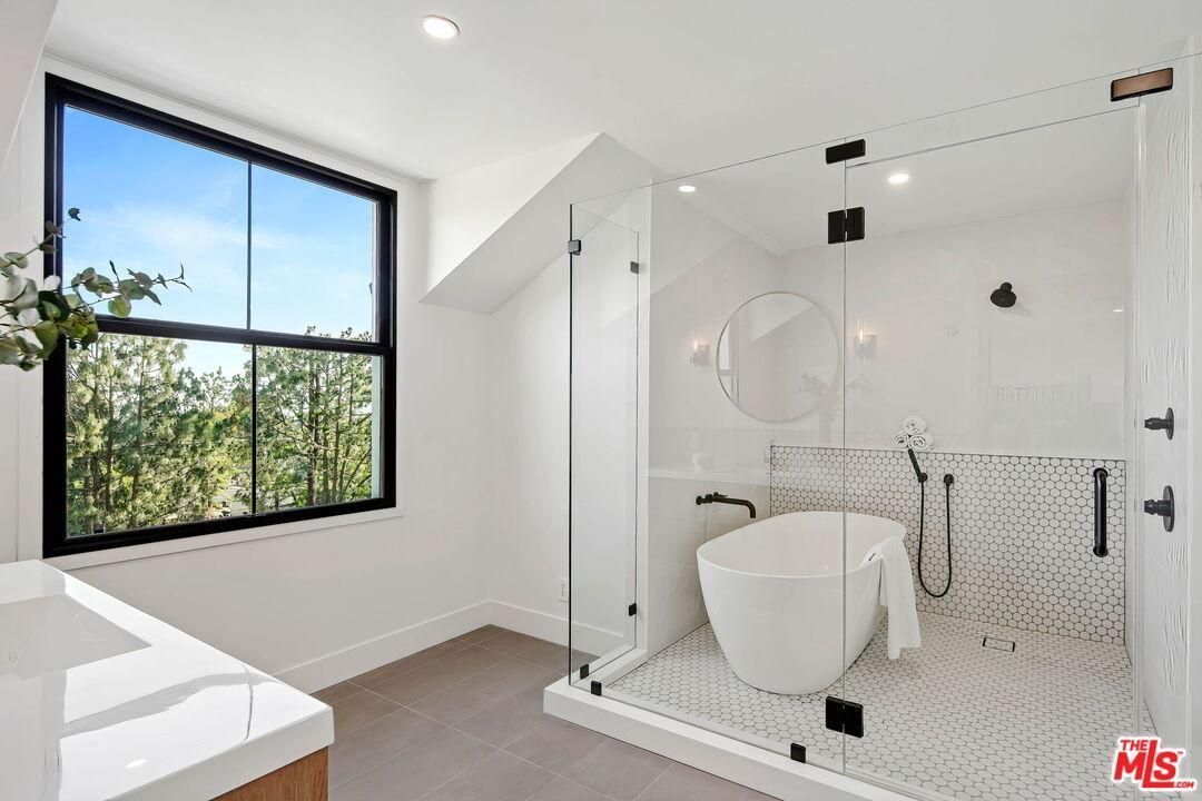 Bright modern bathroom with glass shower, white tub, and large window showing greenery