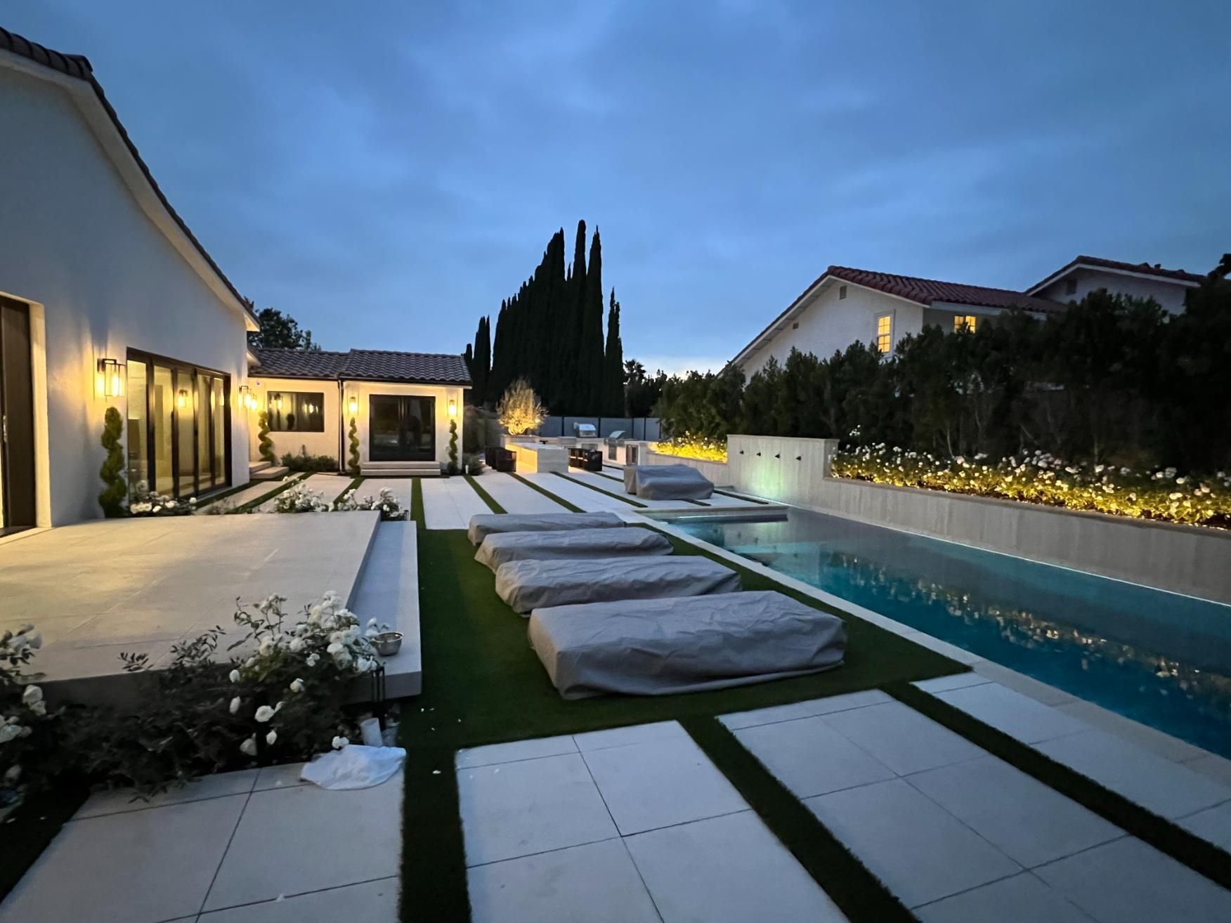 Modern backyard pool at dusk with lounge chairs, white patio seating, and warm house lights