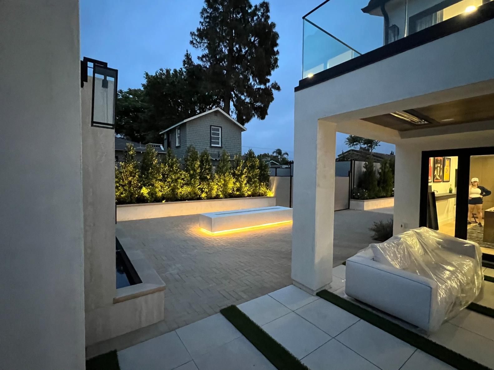 Modern backyard patio at dusk with outdoor seating, lit landscaping, and a two-story house with balcony