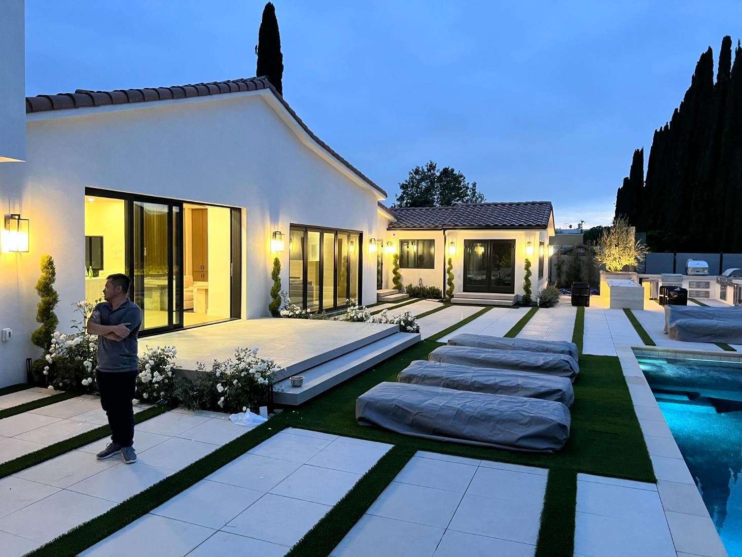Modern home patio at dusk with lit windows, pool, lounge chairs, and a person standing near the house.