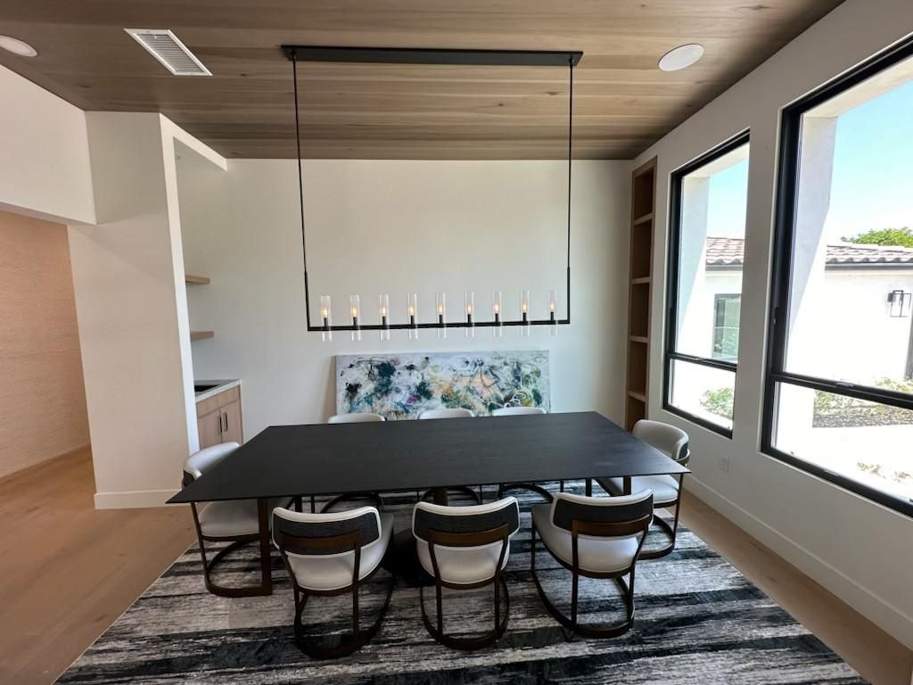 Modern dining room with black table and chairs, patterned rug, large windows, and wood ceiling panel.