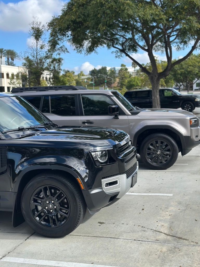 Black and gray Land Rover Defenders parked outdoors under a blue sky, near a tree.