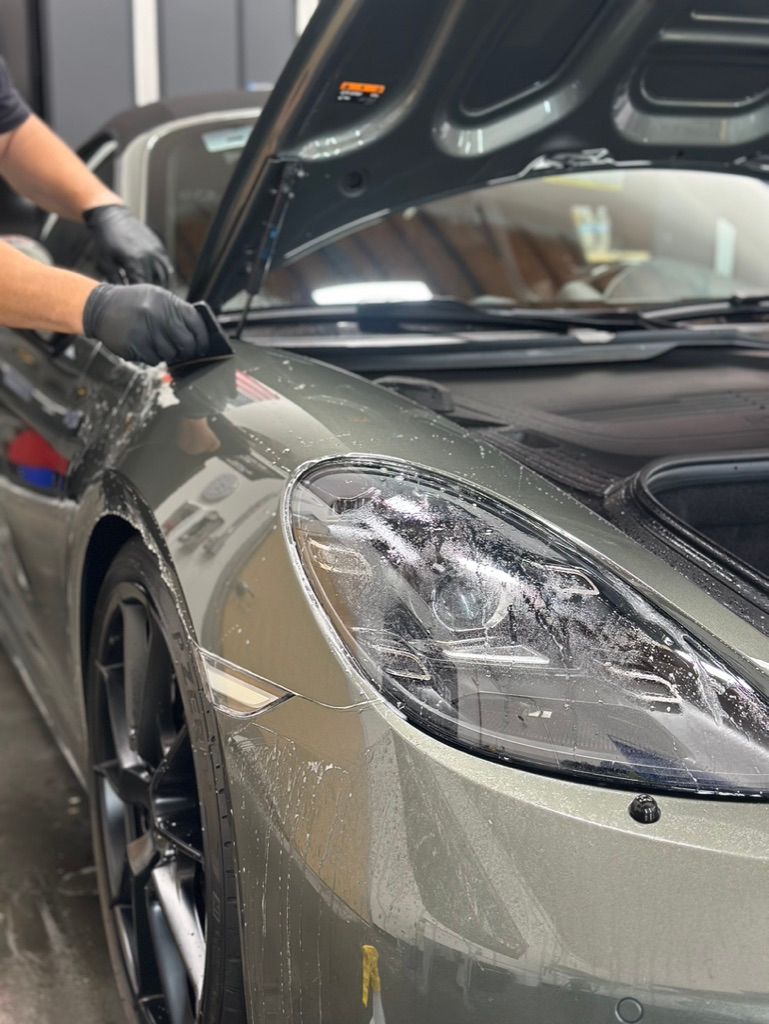 Person applying film to the front fender of a grey sports car, wearing black gloves.