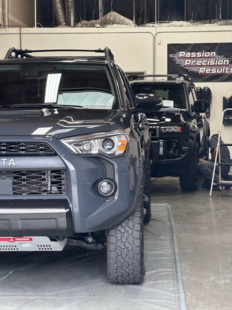 Dark gray Toyota 4Runner with off-road tires, in a shop; a black truck is behind it; a person works beneath it.