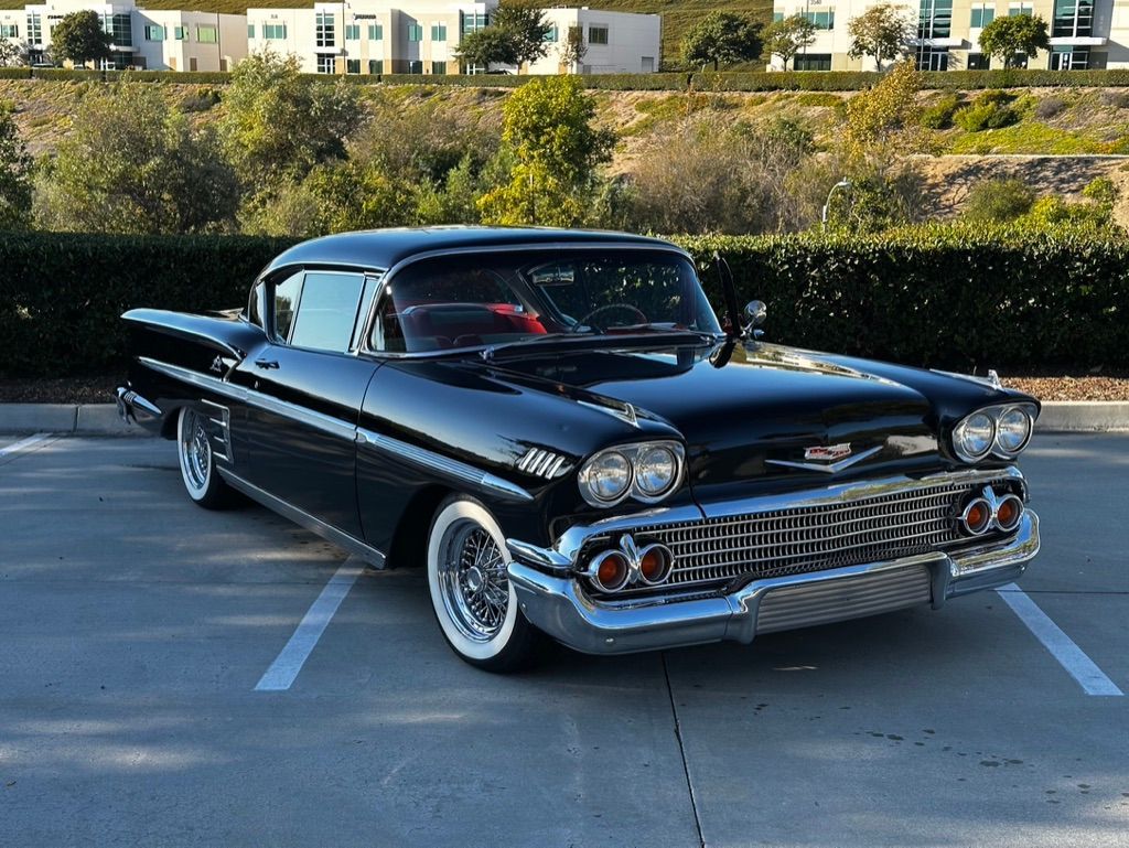 Black 1958 Chevrolet Impala parked in a parking spot with white wall tires and a suburban background.