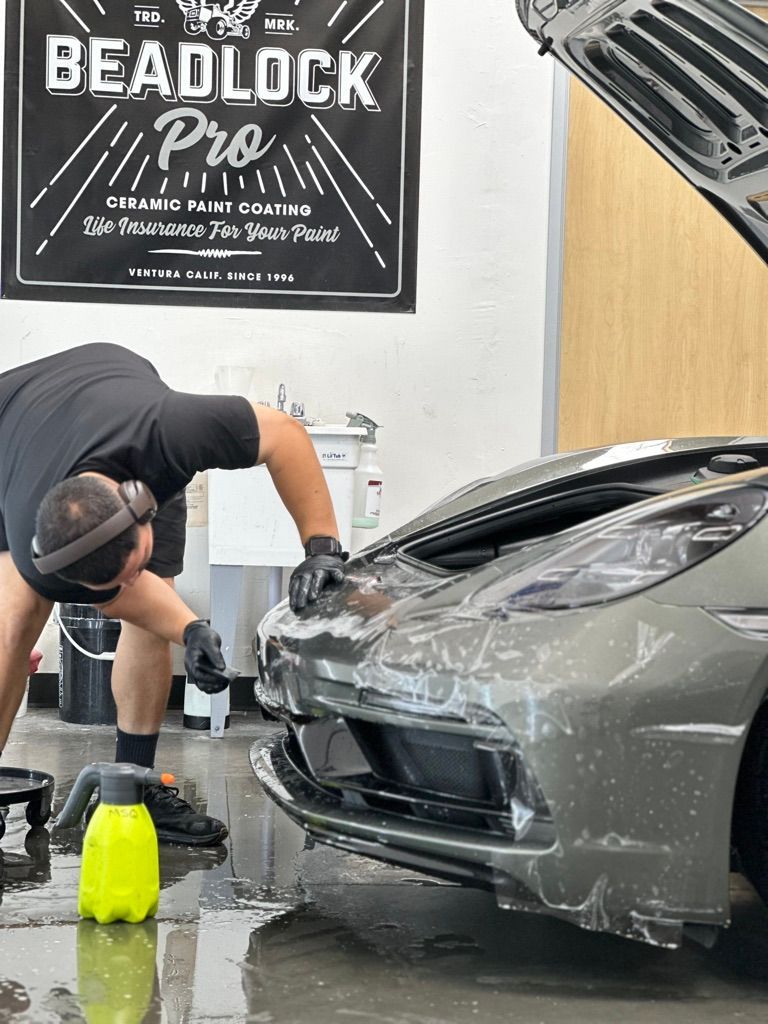 A person washing a gray sports car with a spray bottle in a garage.