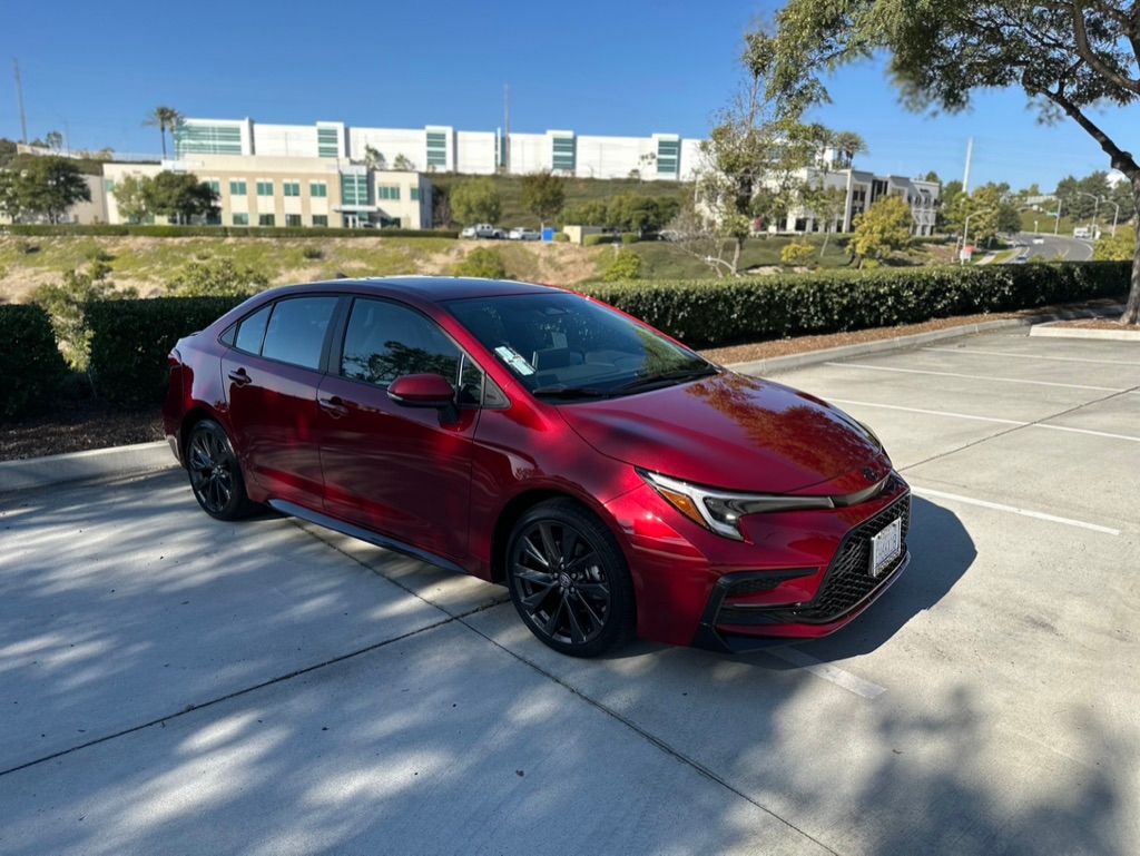 Red Toyota Corolla sedan parked in a lot, with a building and greenery in the background.