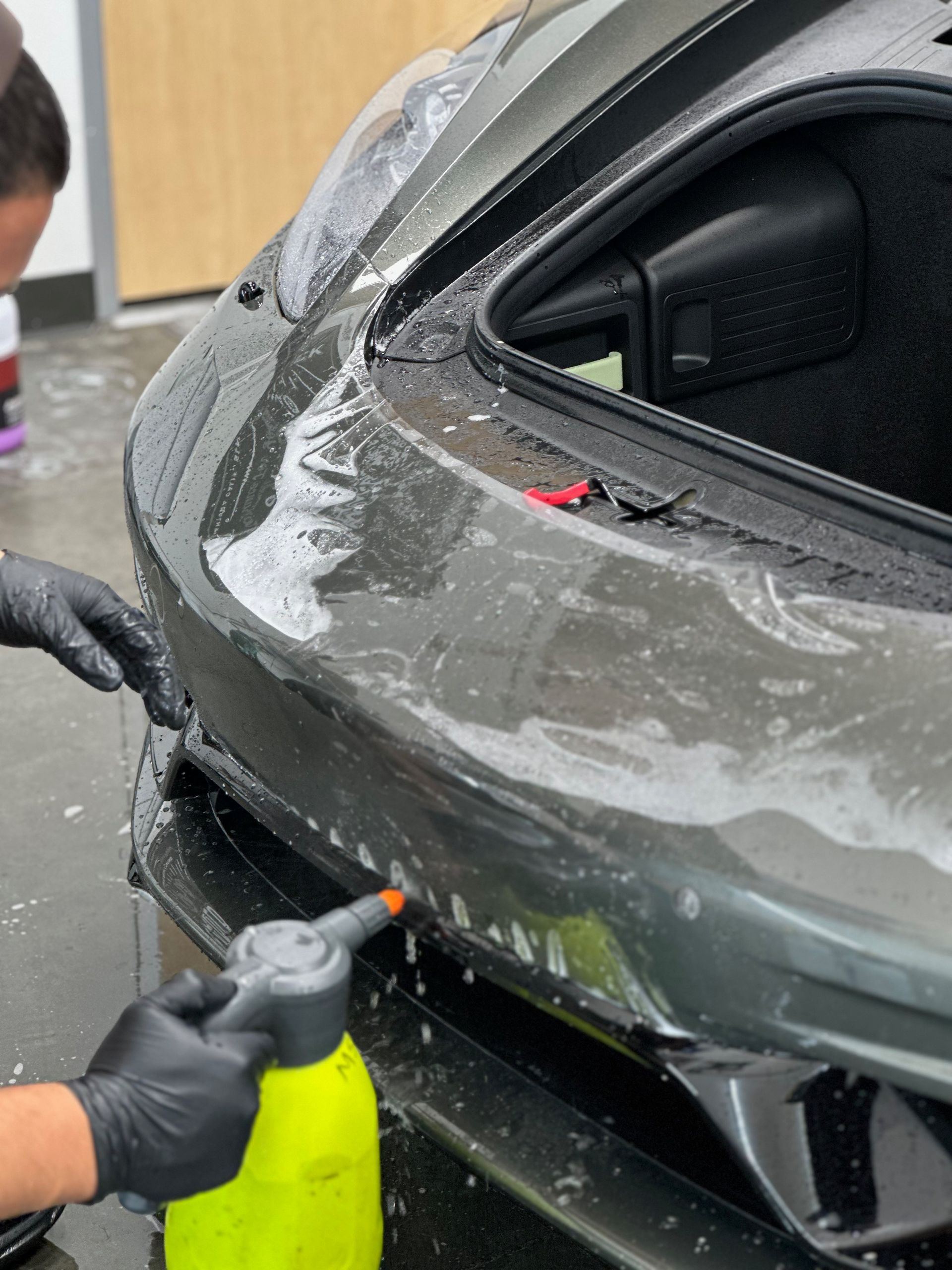 Person applying protective film to a dark gray car bumper, spraying with a yellow bottle.