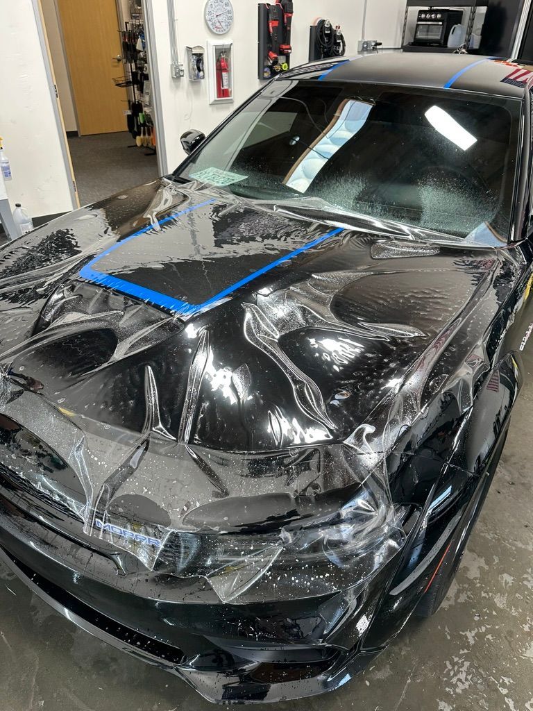 Black car with protective film applied to the hood. Tape is holding the film in place.