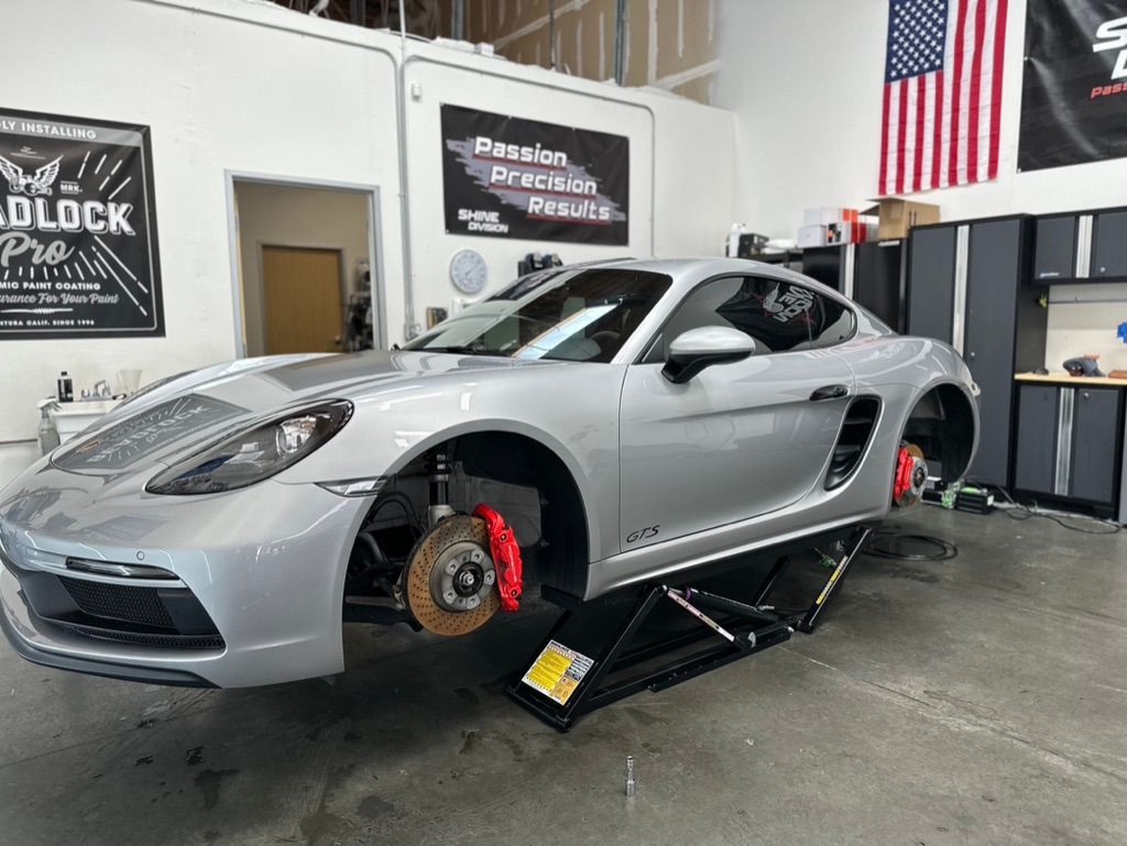 Silver Porsche sports car on a lift in a garage, wheels removed, red brake calipers visible.
