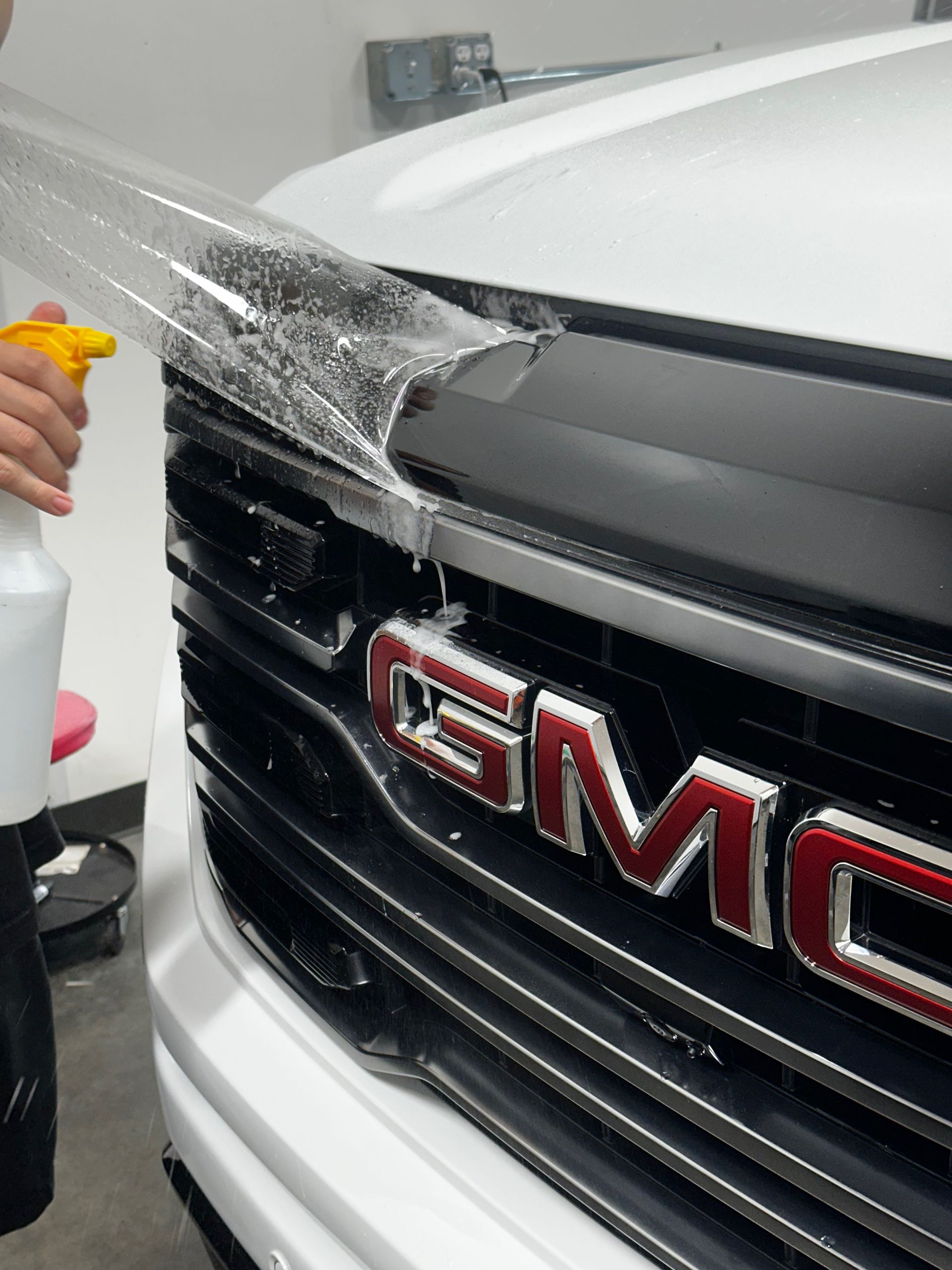 Person spraying water on a clear protective film being applied to a GMC truck grill.