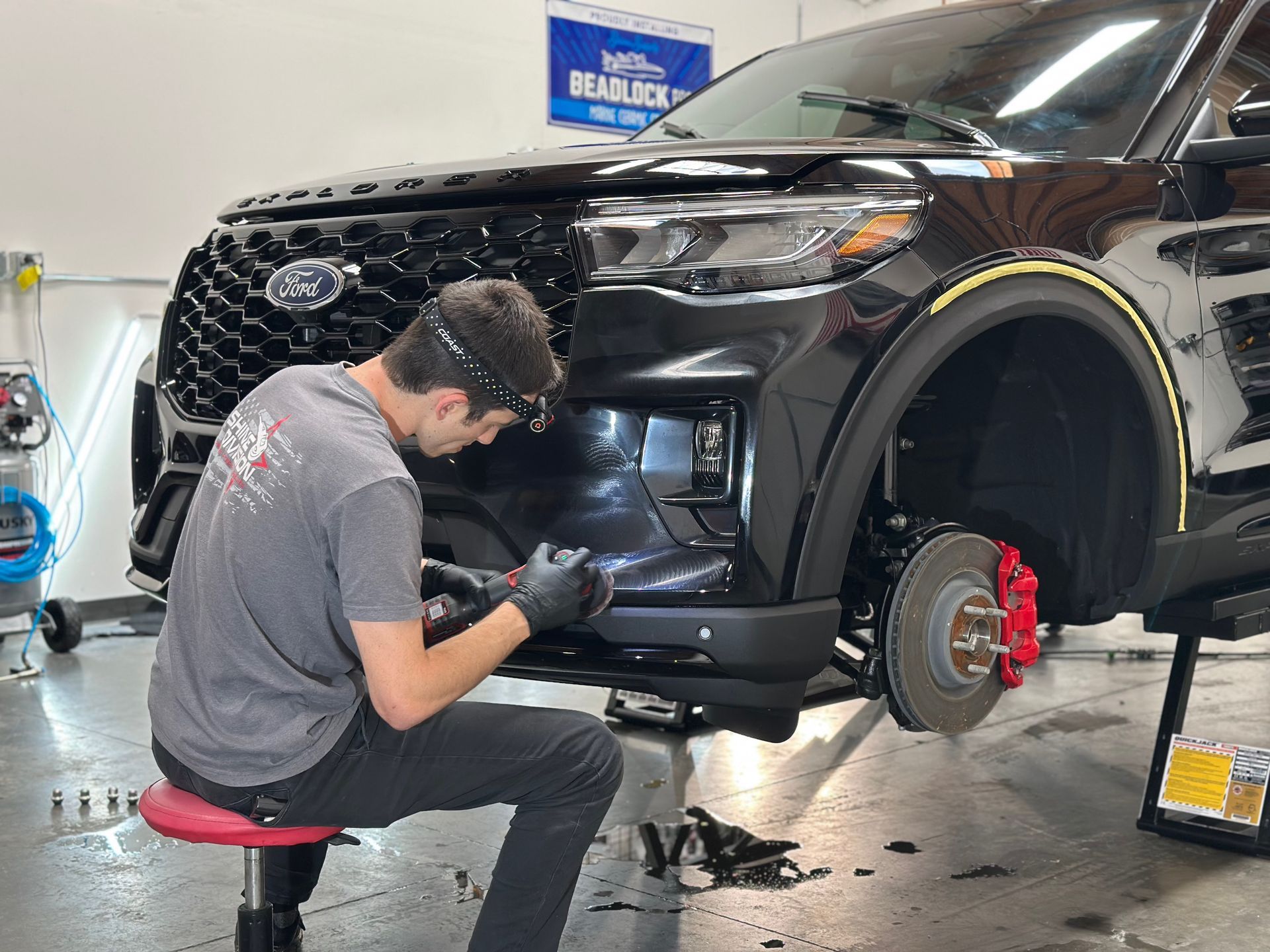 Mechanic working on the front bumper of a black SUV in a shop. Red brake calipers visible.