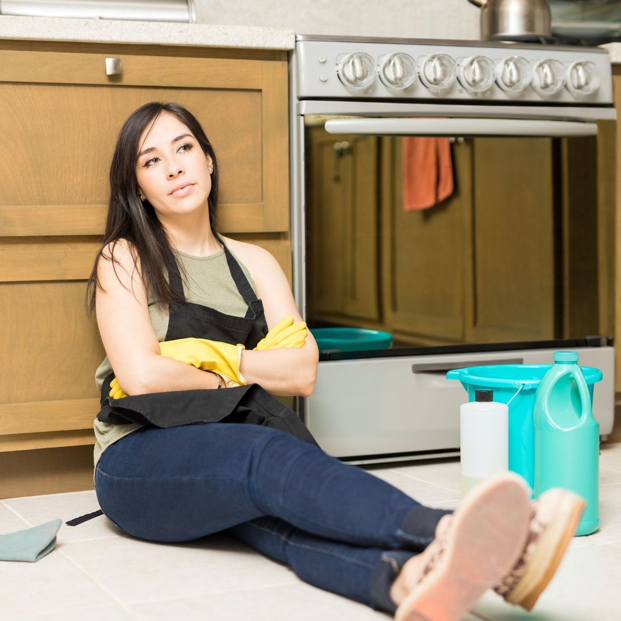 Woman sitting on a kitchen floor with cleaning supplies beside her, arms crossed and looking at the camera.