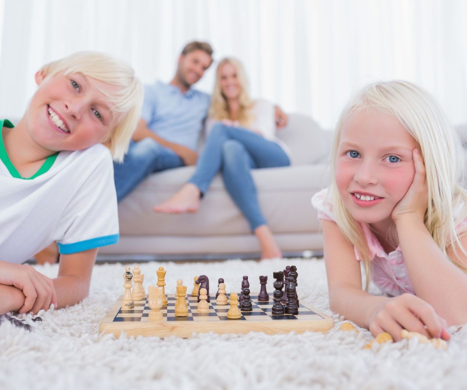 Children playing chess on a carpet while two adults sit smiling on a couch in the background
