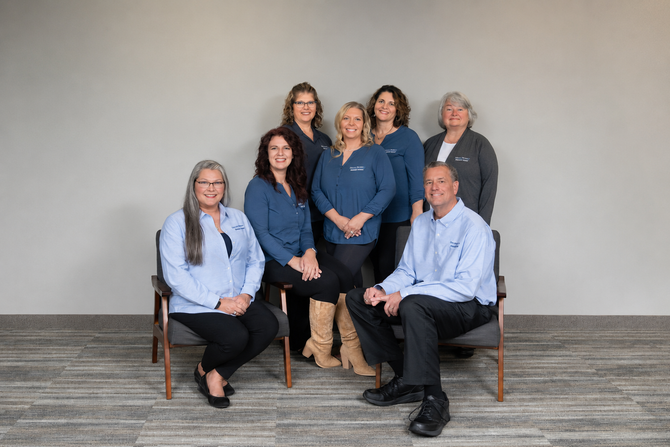 Seven people posed in a group portrait against a plain gray wall, seated and standing in business attire.