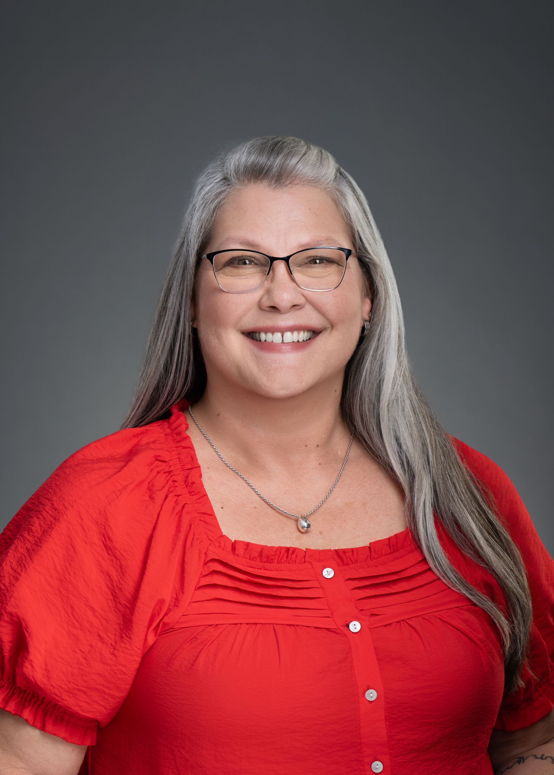Smiling person with long gray hair in a blue button-up shirt against a gray studio backdrop