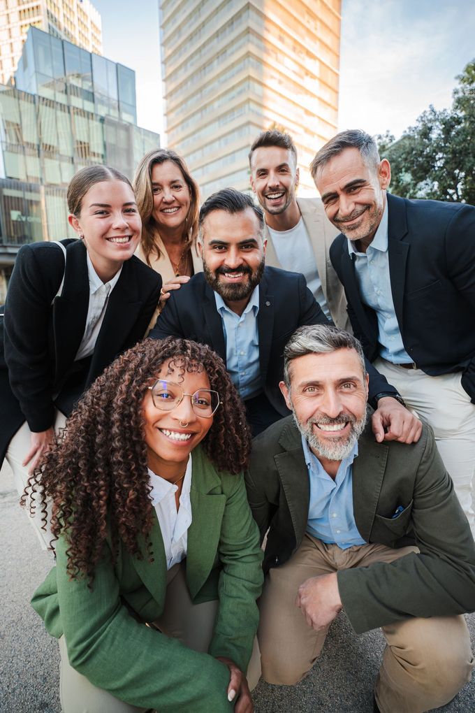 Group of six smiling coworkers posing outdoors in business attire in front of modern office buildings