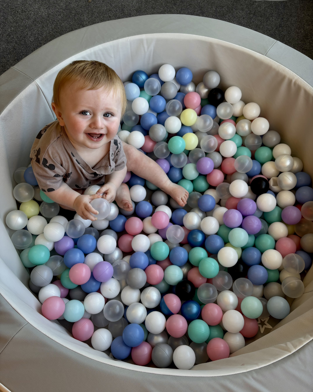 Child sitting in a ball pit filled with pastel-colored balls, smiling up at the camera