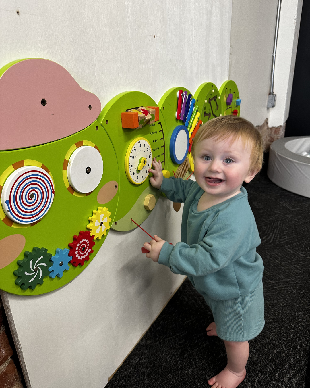 Toddler touching a colorful wall activity panel and smiling in a playroom.