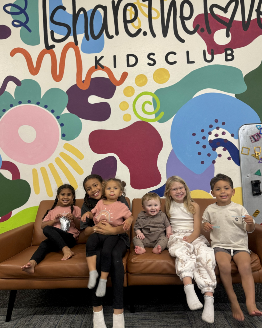 Children sitting on a bench in front of a colorful Kids Club mural
