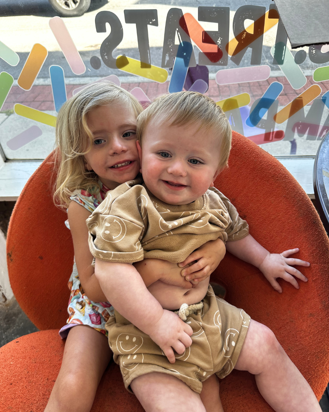 Two smiling children sitting together on an orange chair by a decorated window