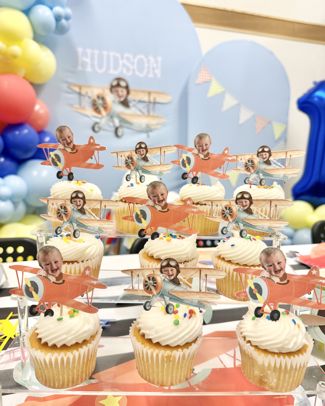 Birthday dessert table with cupcakes, blue-and-yellow balloon decor, and airplane-themed photo toppers