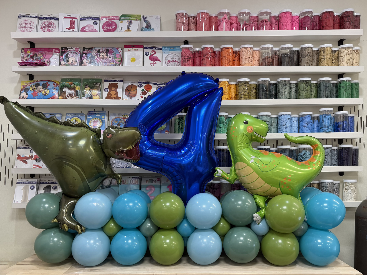 Balloon display with metallic letters and a green dinosaur balloon in front of colorful balloon stacks and shelves.