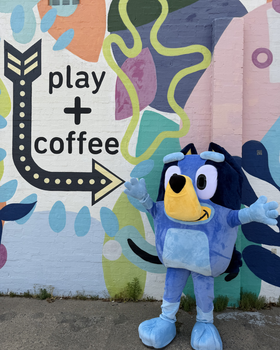 Blue cartoon dog mascot posing by a mural with “play + coffee” text and an arrow.