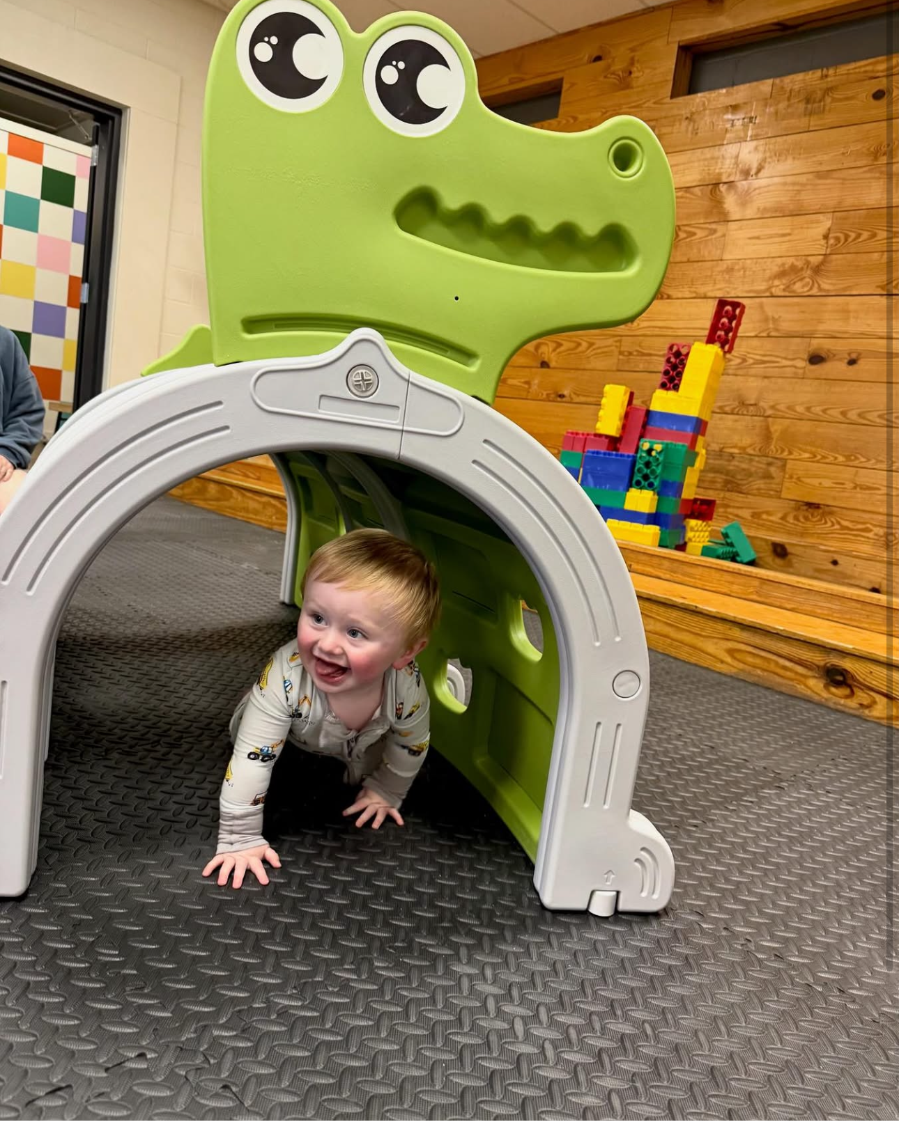 Smiling toddler crawling through a white arch tunnel in a playroom with a green crocodile cutout and blocks