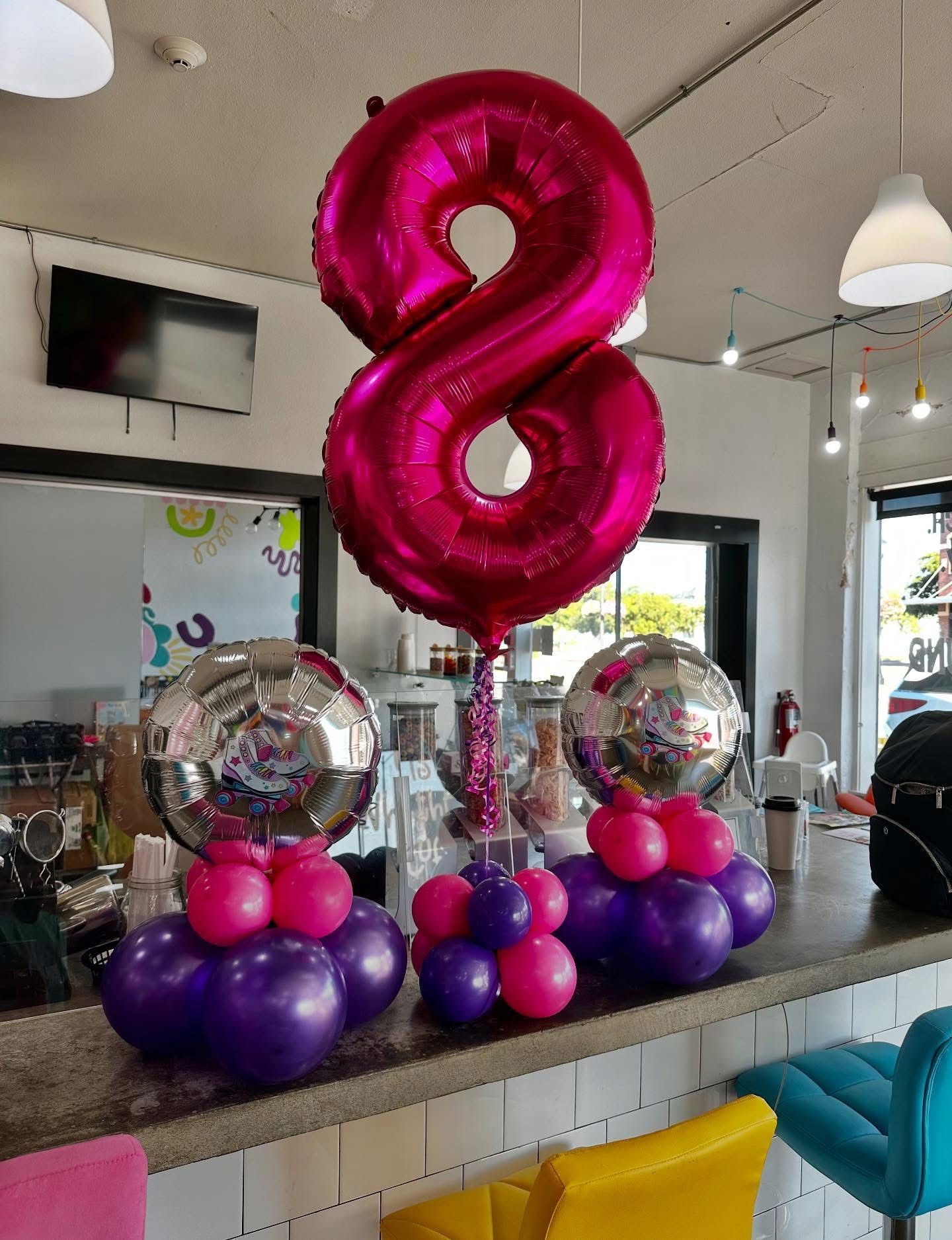 Pink number 8 balloon centerpiece with purple and pink balloon clusters on a counter in a party venue