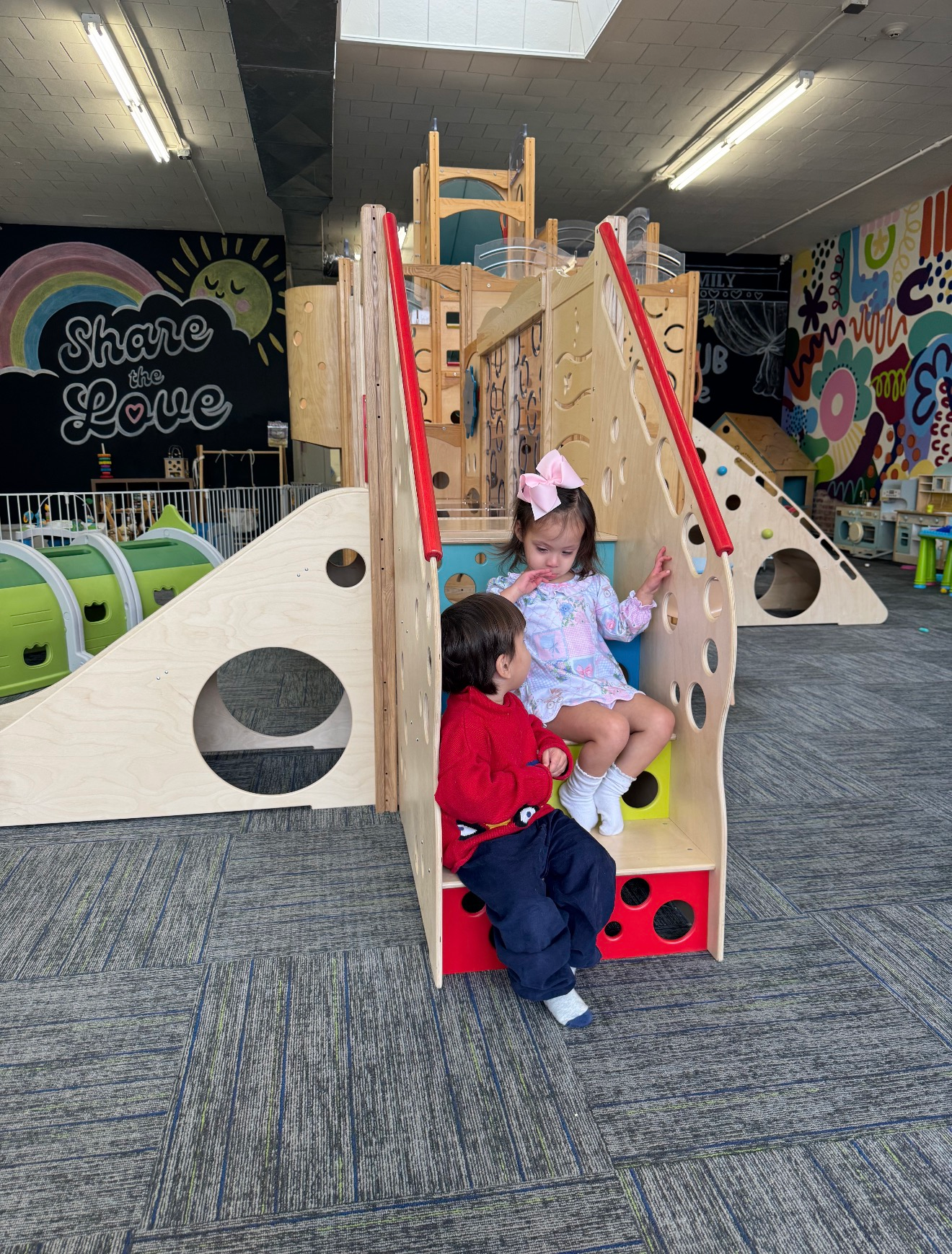 Two children sit on a colorful indoor playground climbing structure with slides and cutout windows.