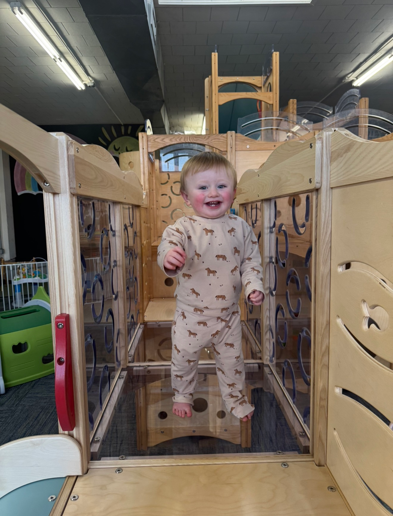 Smiling toddler standing on a wooden indoor play structure, reaching toward the camera.