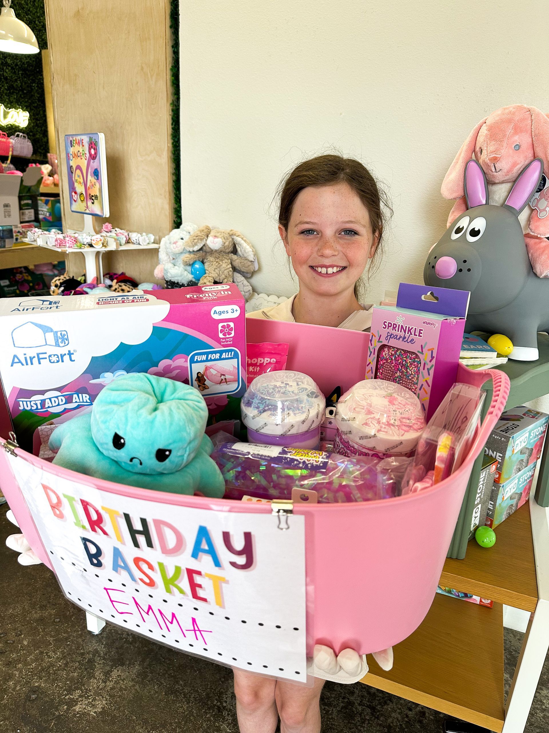 Child behind a pink birthday basket filled with gifts, smiling in a decorated room.