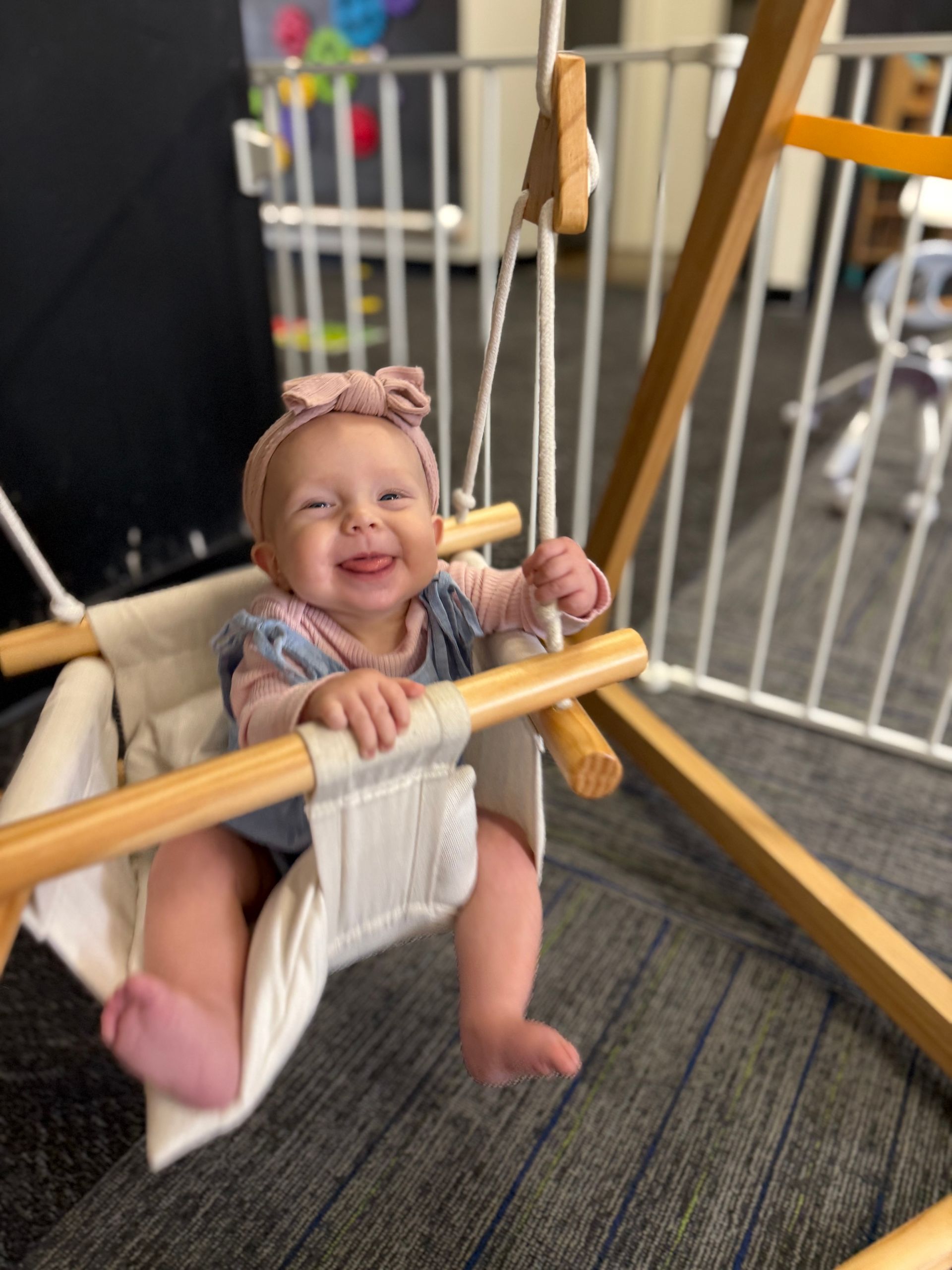 Smiling baby in a white swing indoors, holding the ropes and wearing a pink bow headband.