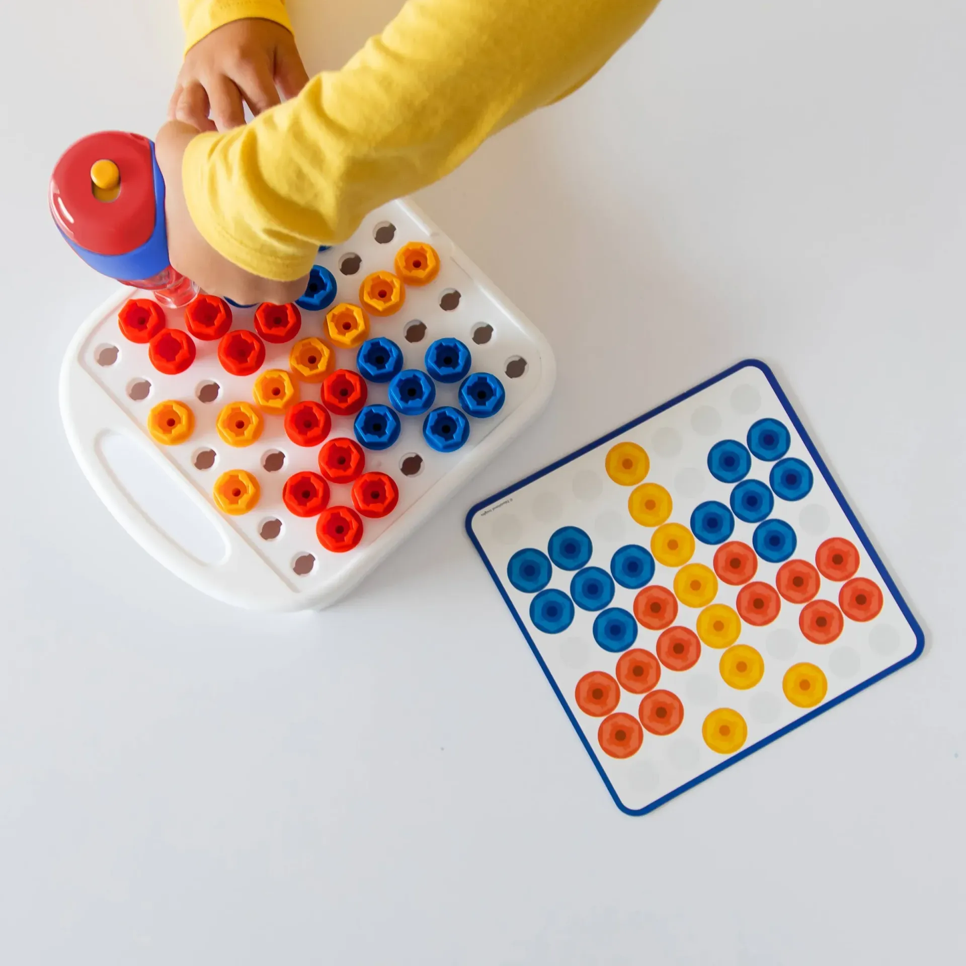 Hands placing colorful pegs on a pegboard next to a pattern card on a white table