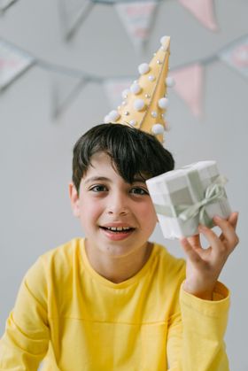 Child in yellow shirt holding a wrapped gift and wearing a party hat, smiling indoors.