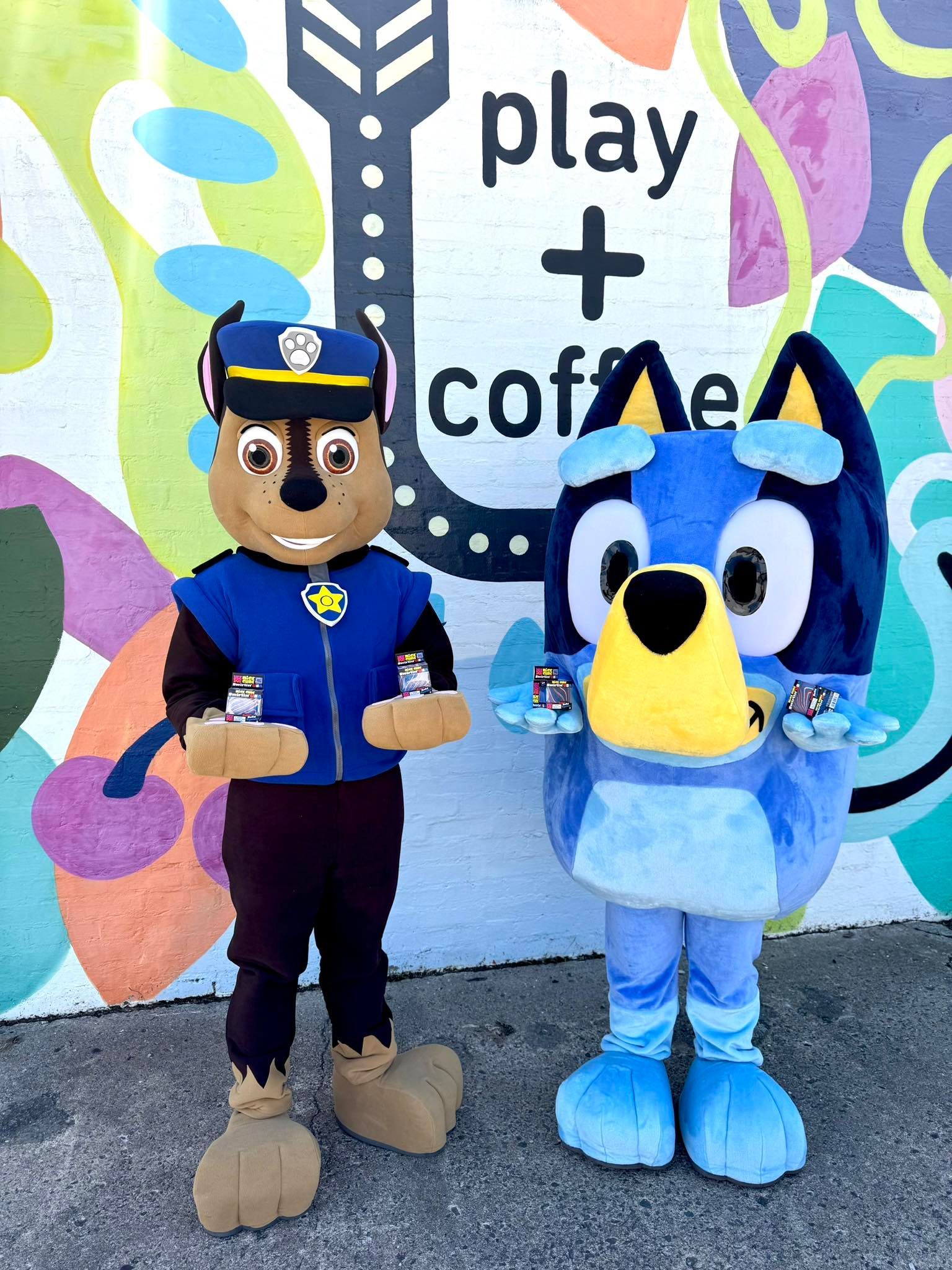 Two mascots in blue uniforms posing in front of a colorful “play + coffee” mural.