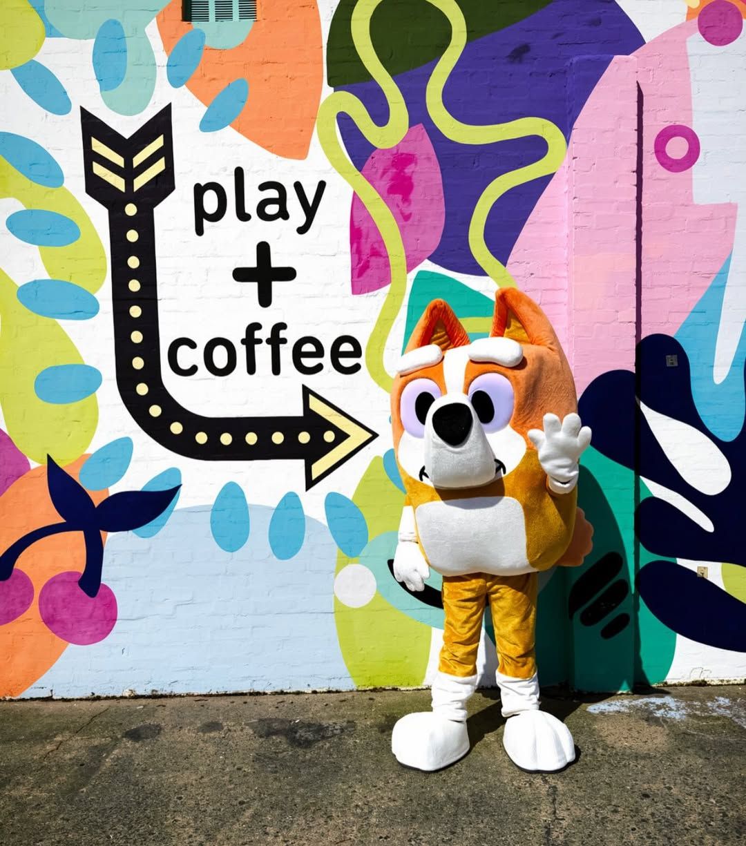 Mascot in orange and white stands by colorful mural reading “play + coffee” with an arrow.