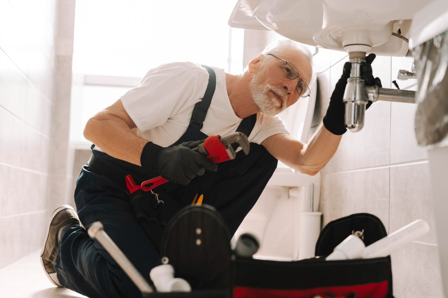 Plumber working under a bathroom sink, holding a wrench, wearing black gloves, and looking up.