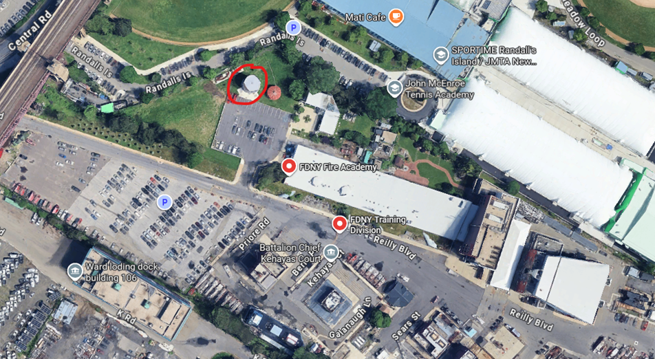 Satellite view of a parking lot, green space with a circular structure, and surrounding buildings in an urban area.