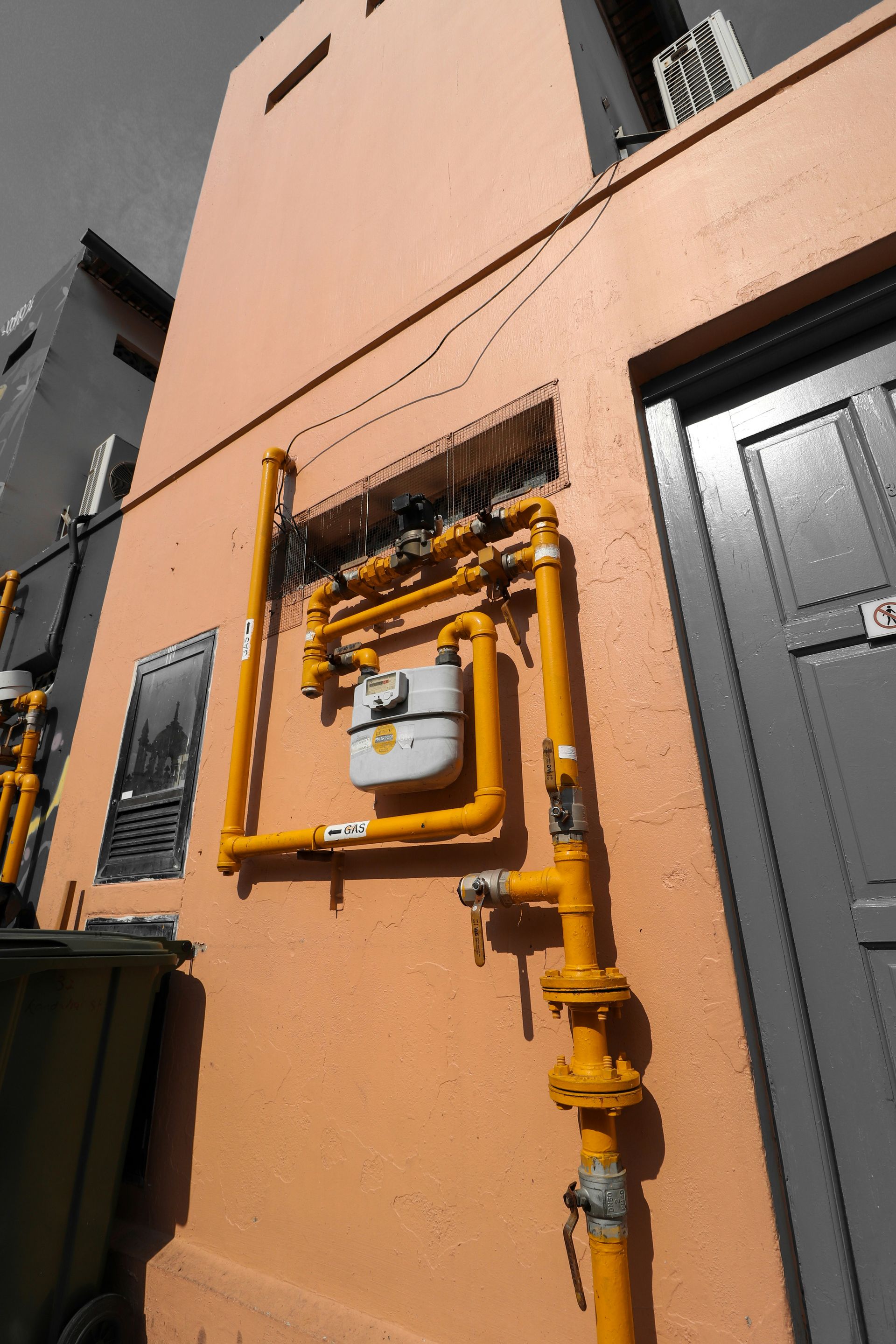 Exterior wall with yellow gas pipes, meter, and door. Green trash can on left. Orange wall.